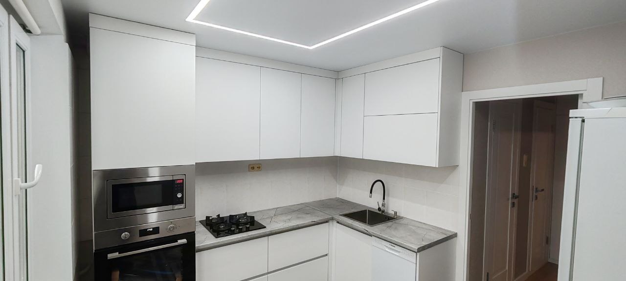Nowoczesna kuchnia z białymi, minimalistycznymi szafkami, blatem imitującym marmur, wbudowanym piekarnikiem i mikrofalówką ze stali nierdzewnej oraz oświetleniem LED w suficie.
