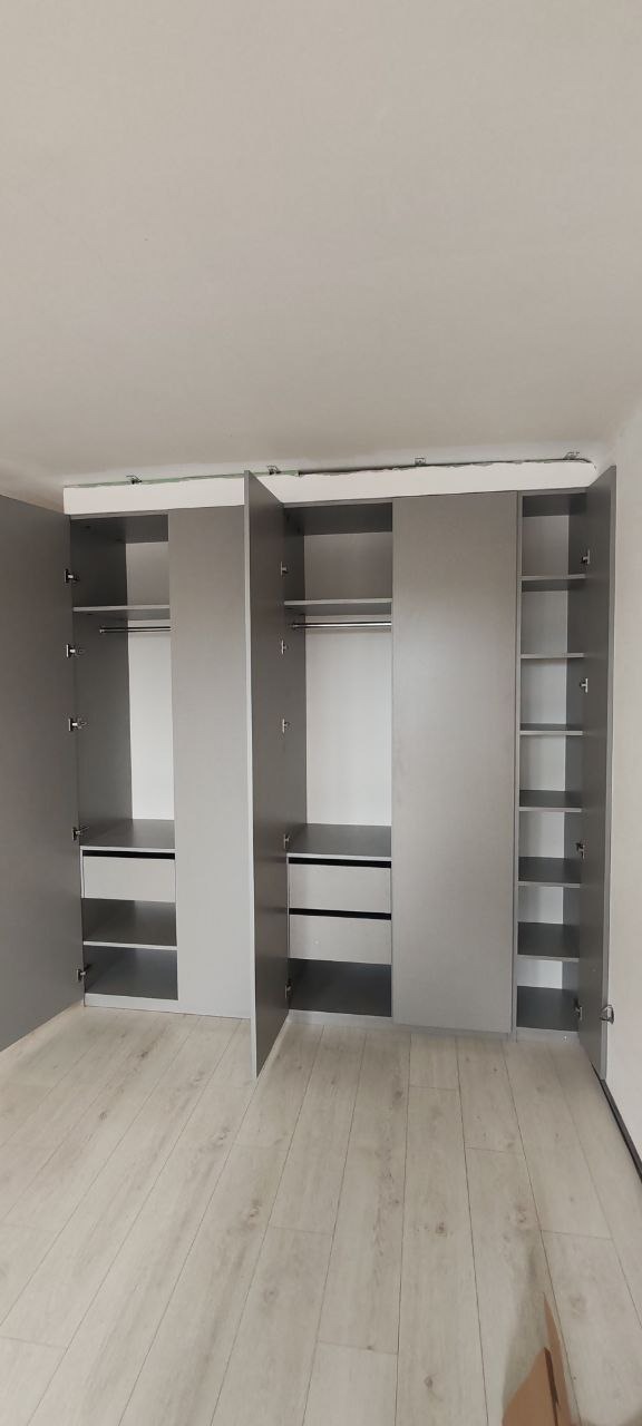 Szara, trzydrzwiowa szafa wnękowa z otwartymi drzwiami, ukazująca półki i drążki na ubrania, zamontowana w jasnym pomieszczeniu z panelami podłogowymi.