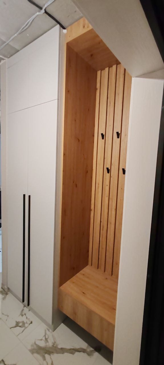 Biała szafa z długimi, czarnymi uchwytami sąsiadująca z drewnianą wnęką z wieszakami na ubrania i siedziskiem, całość na marmurowej podłodze.