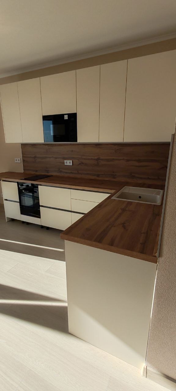 Nowoczesna kuchnia z białymi szafkami i blatem imitującym drewno, z wbudowanym piekarnikiem i płytą grzewczą oraz zlewozmywakiem w rogu.