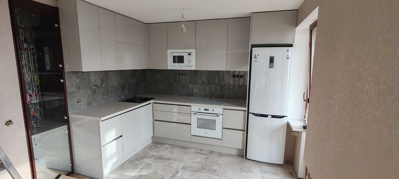 Nowoczesna kuchnia z błyszczącymi, szarymi szafkami, ciemnoszarymi płytkami nad blatem i białym sprzętem AGD, w tym lodówką i kuchenką, oświetlona wiszącą lampą.