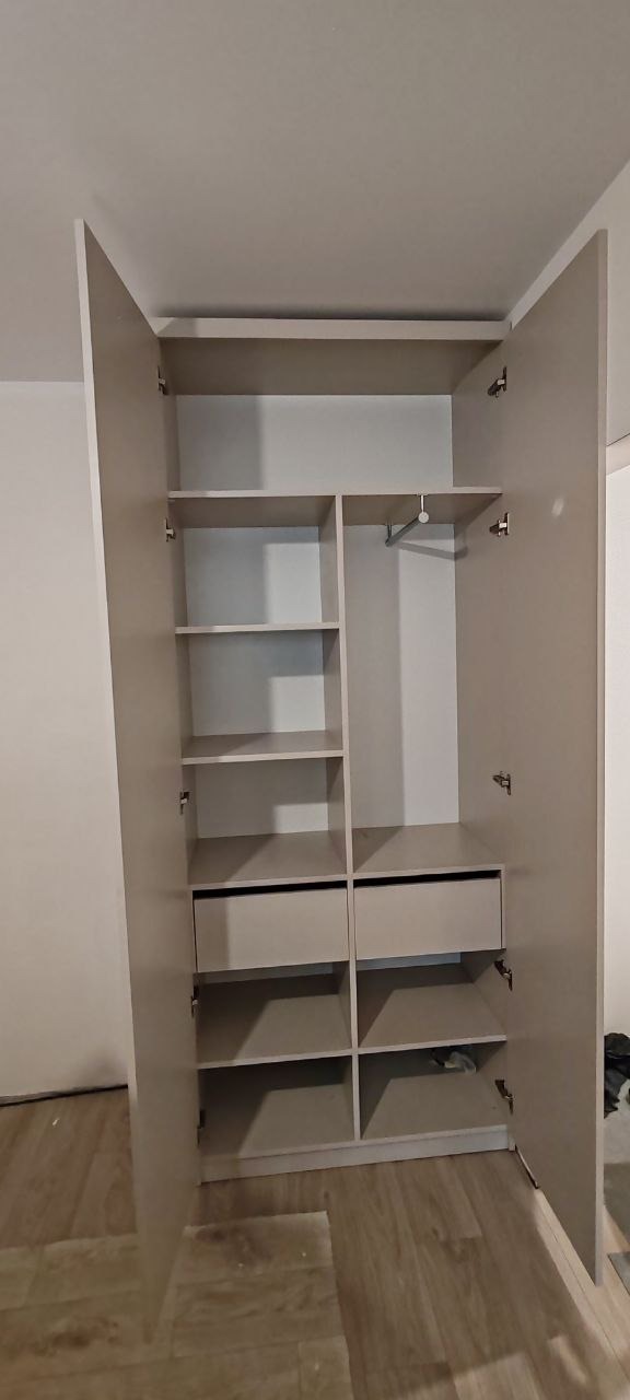 Wnętrze szafy wnękowej z otwartymi drzwiami, widoczne półki, szuflady i drążek na ubrania, neutralna kolorystyka.