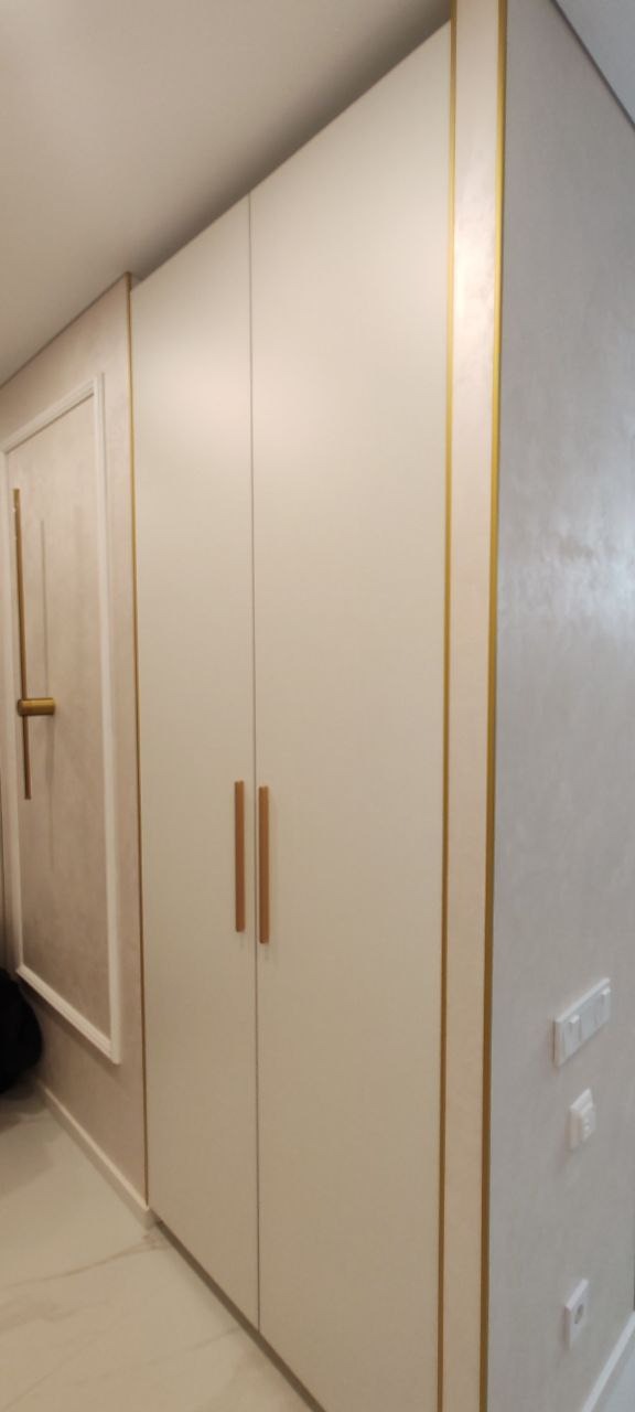 Biała szafa wnękowa z drewnianymi uchwytami i złotymi akcentami wykończeniowymi, dopasowana do koloru ramy drzwi w tle i dekoracyjnej ściany ze srebrzystą fakturą.