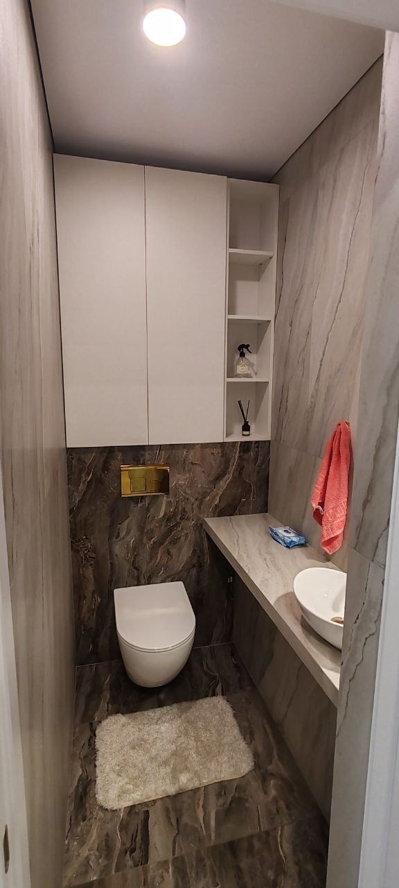 Nowoczesna toaleta w odcieniach szarości i brązu z wiszącą muszlą, umywalką nablatową i szafką wiszącą z otwartymi półkami, uzupełniona o miękki dywanik i akcesoria.