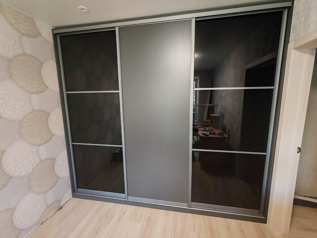 Trzydrzwiowa szafa przesuwna z ciemnymi lustrzanymi drzwiami i szarymi panelami, widoczna na tle jasnej ściany w okrągłe wzory i drewnianej podłogi.