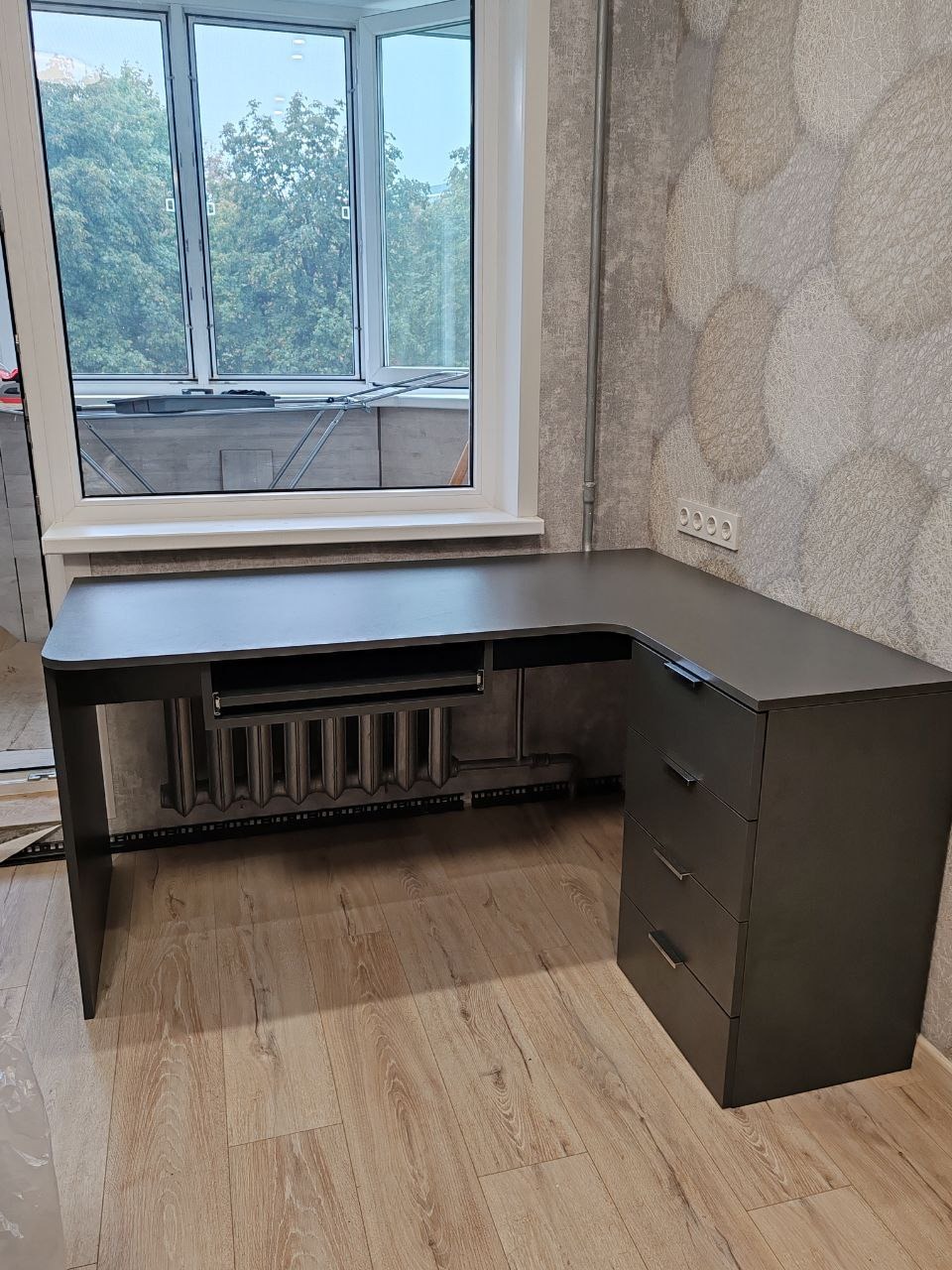 Ciemnoszare biurko narożne z trzema szufladami, zamontowane w pokoju w Warszawie, z widokiem na balkon i drzewa za oknem.