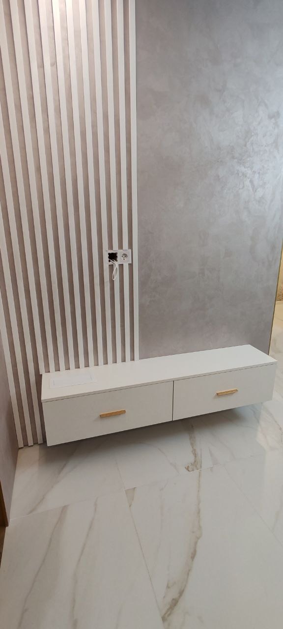 Nowoczesna biała szafka RTV z dwoma szufladami z poziomymi, złotymi uchwytami, zamontowana na ścianie z dekoracyjnymi pionowymi listwami i szarą, teksturowaną ścianą w tle; widoczny fragment...