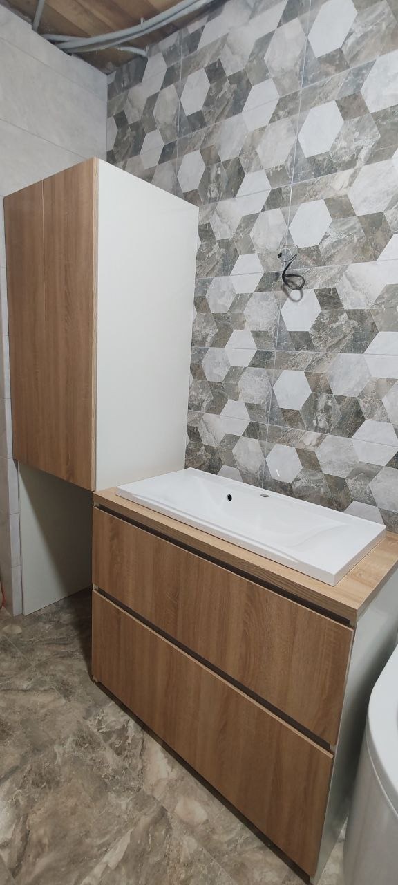 Nowoczesna łazienka z szafką pod umywalkę z drewnopodobnymi frontami, białą umywalką nablatową i wiszącą szafką. Na ścianie dekoracyjne płytki w geometryczne wzory.