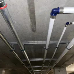 Równolegle prowadzone rury instalacyjne pod betonowym stropem, mocowane za pomocą uchwytów do szyn montażowych; widoczne rury stalowe i z tworzywa sztucznego z niebieskimi złączkami.