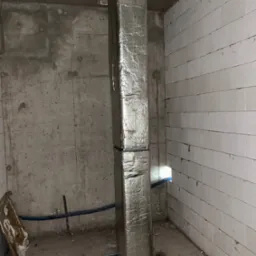 Pionowy kanał wentylacyjny owinięty folią izolacyjną, zamontowany w surowym pomieszczeniu z betonowymi i bloczkowymi ścianami, widoczne elementy instalacji i resztki materiałów budowlanych.