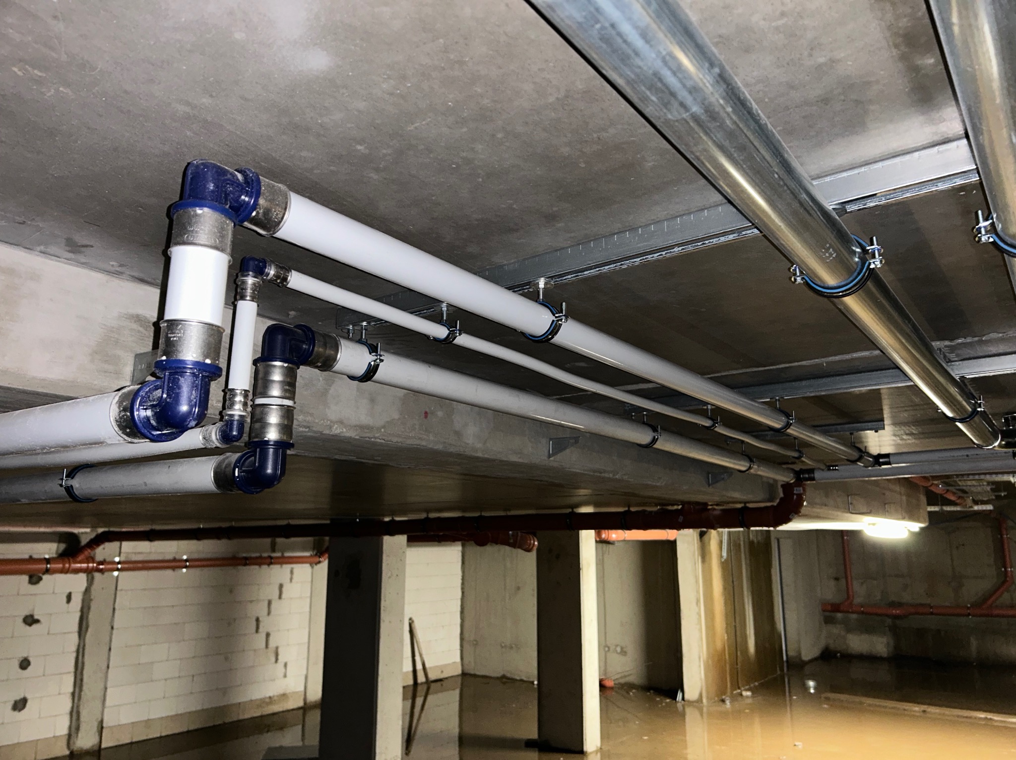 Instalacja hydrauliczna pod stropem w zalewanym pomieszczeniu w Wąsoszu, widoczne białe i szare rury z niebieskimi złączkami.