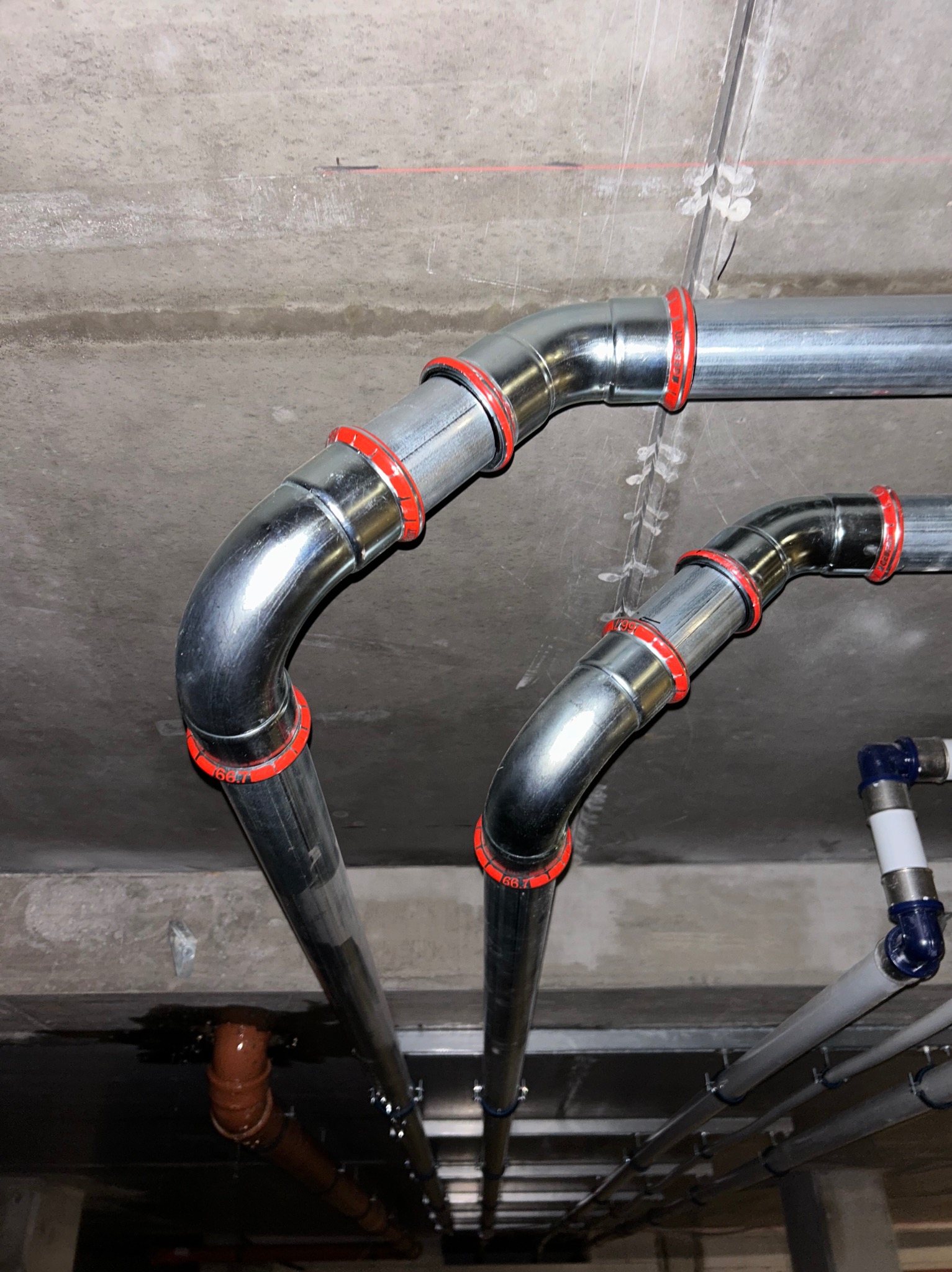 Metalowe rury wentylacyjne z czerwonymi uszczelnieniami, zamontowane pod betonowym sufitem z widocznymi elementami mocującymi i poziomą linią lasera.