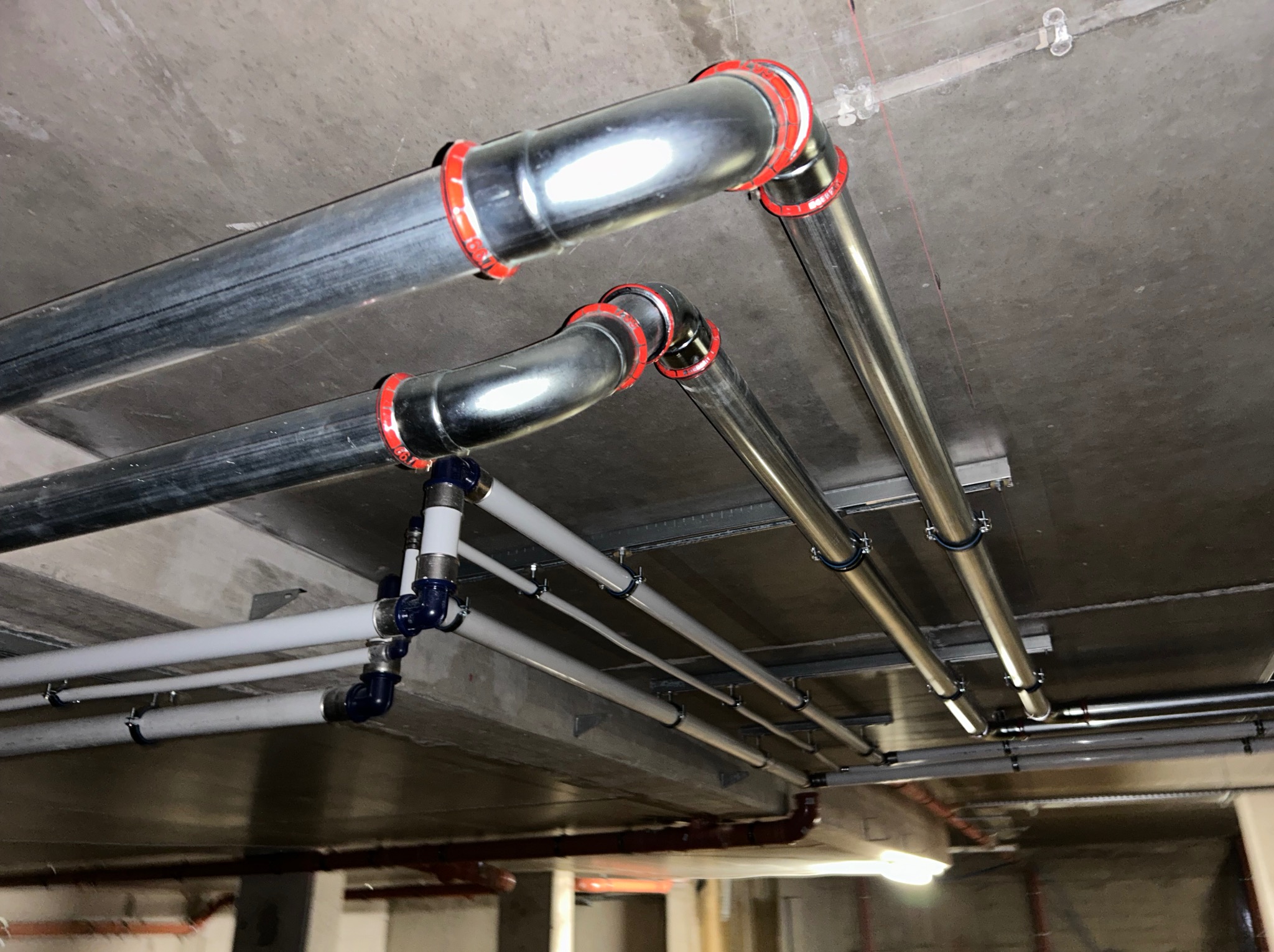 Różne rodzaje rur instalacyjnych pod sufitem, w tym metalowe rury wentylacyjne z czerwonymi uszczelkami i białe rury z niebieskimi złączkami, zamocowane do betonowego sufitu.