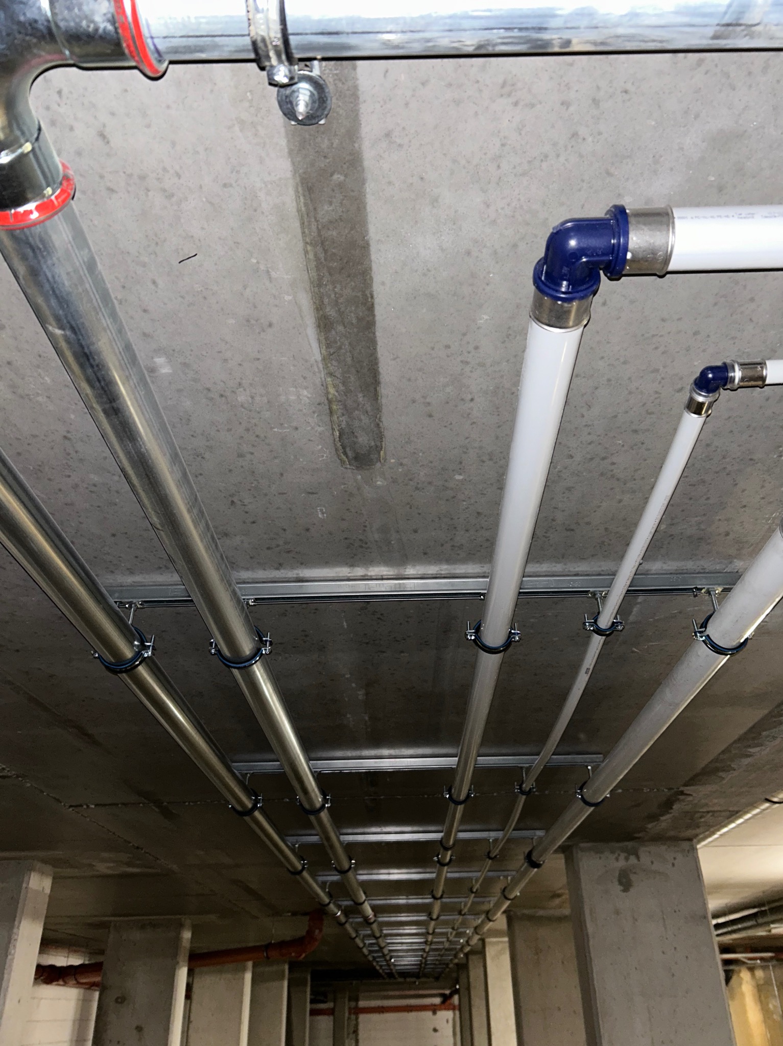 Równolegle prowadzone rury instalacyjne pod betonowym stropem, mocowane za pomocą uchwytów do szyn montażowych; widoczne rury stalowe i z tworzywa sztucznego z niebieskimi złączkami.