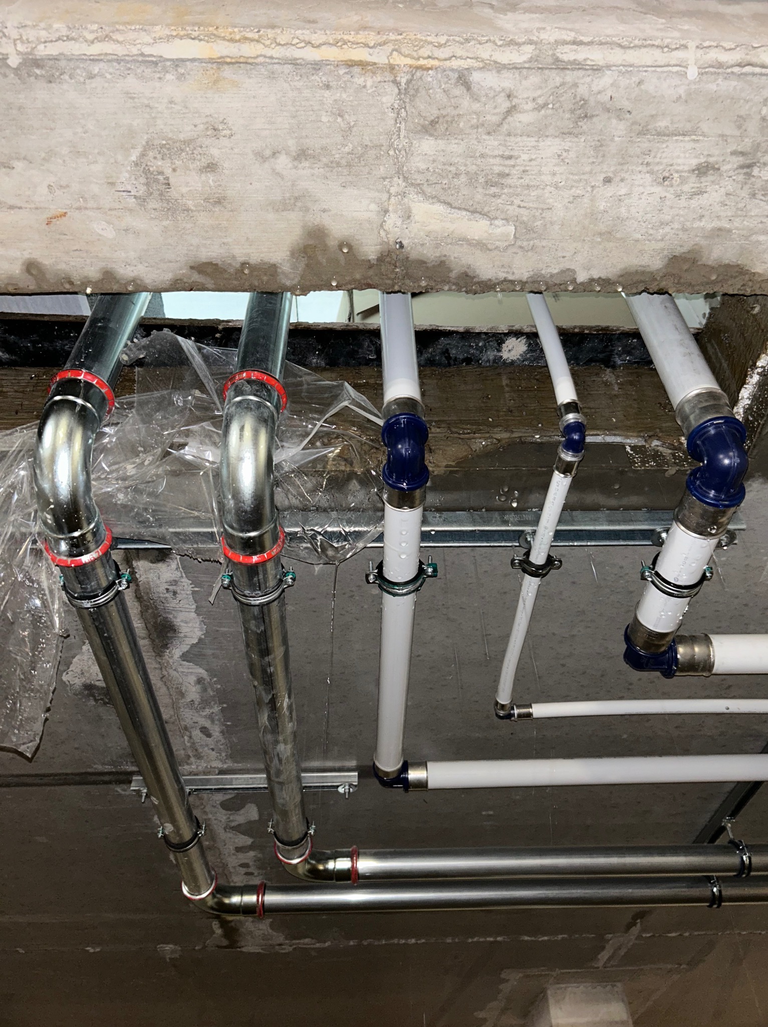 Montaż instalacji z rur stalowych i plastikowych pod betonowym sufitem, widoczne złącza i uchwyty mocujące, folia ochronna na elementach konstrukcyjnych.