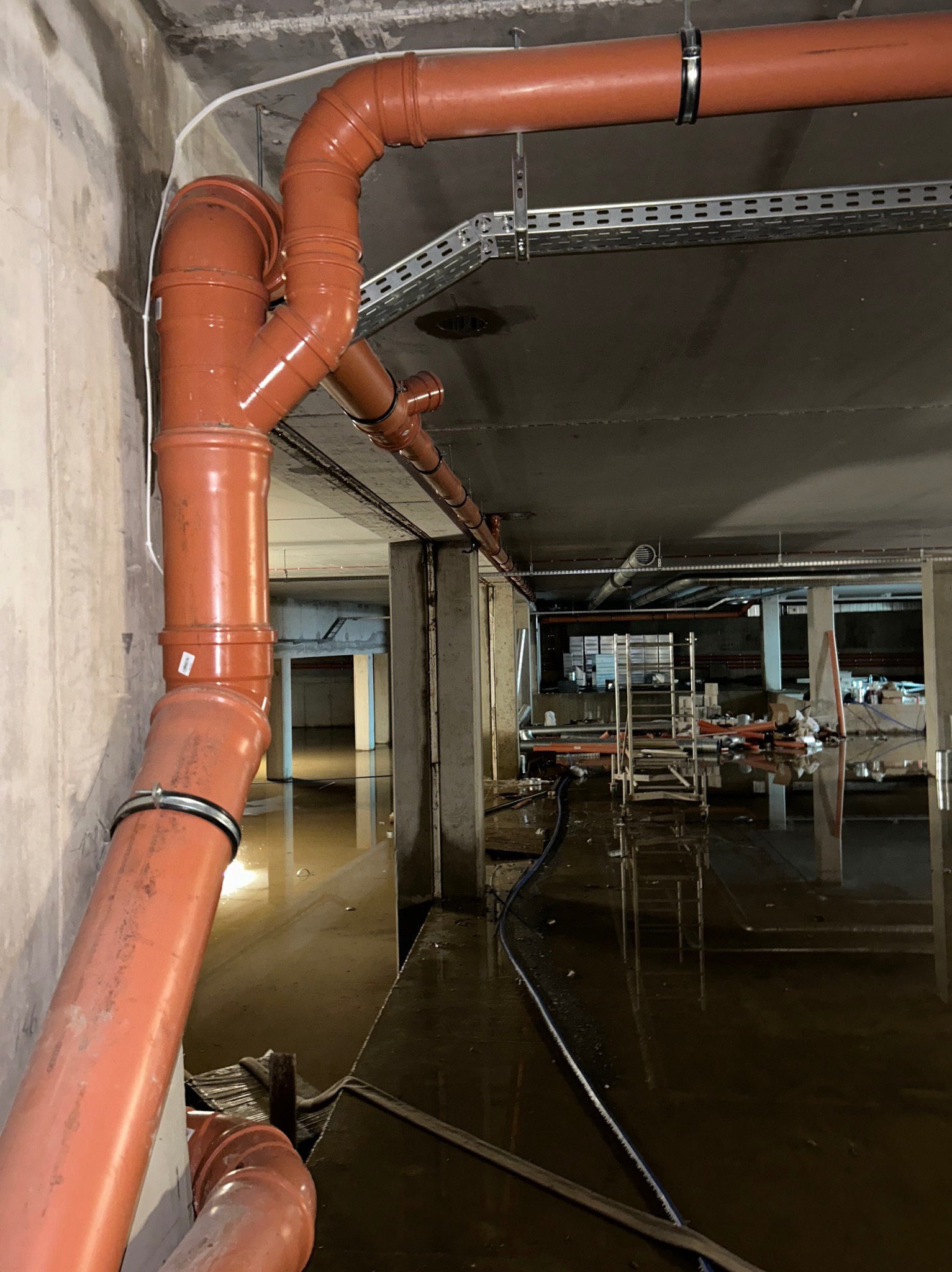 Montaż pomarańczowych rur kanalizacyjnych z obejmami w surowym wnętrzu budynku, częściowo zalane wodą, widoczne elementy konstrukcyjne i rusztowania.