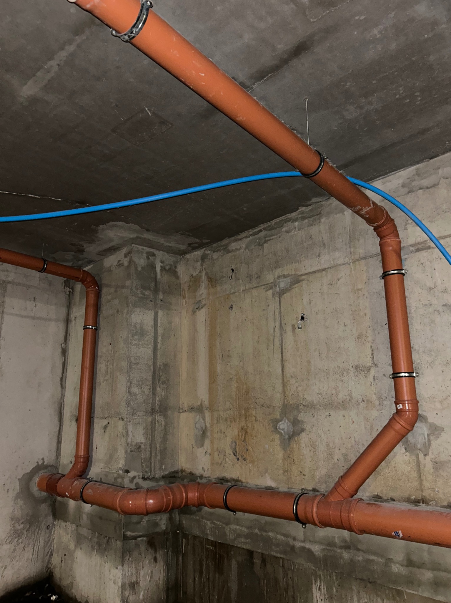 Pomarańczowe rury kanalizacyjne z czarnymi obejmami, zamontowane pod betonowym sufitem w surowym wnętrzu. Dodatkowo widoczna niebieska rurka.