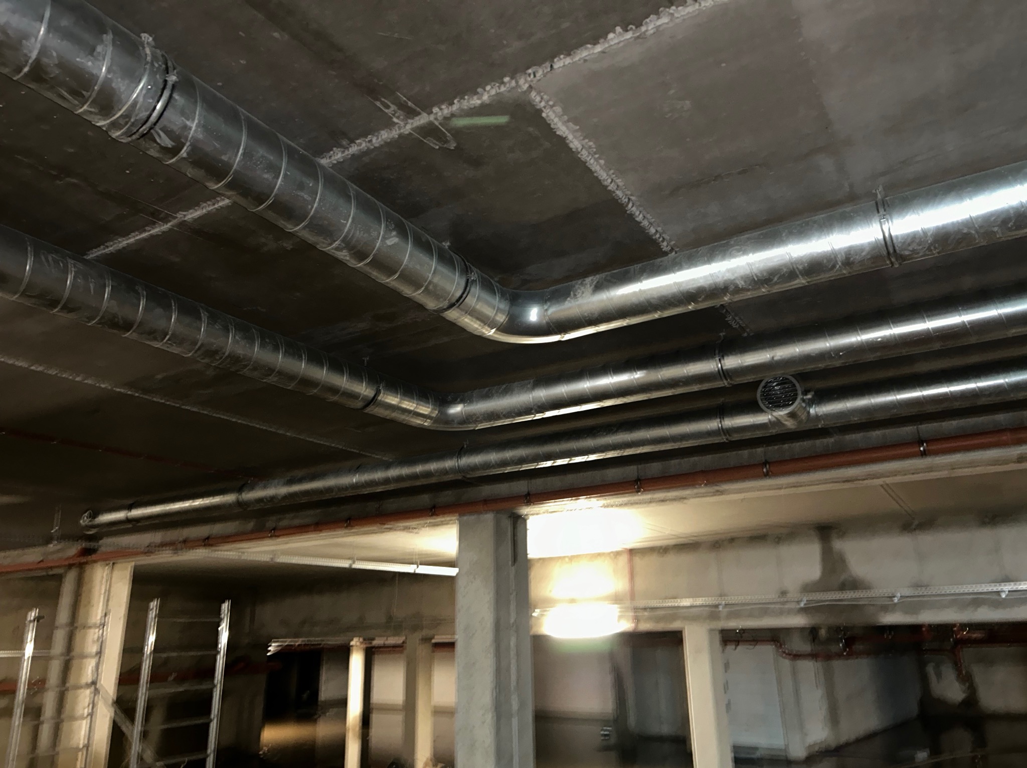 Metalowe rury wentylacyjne spiralne pod betonowym sufitem w surowym wnętrzu budynku w trakcie budowy, widoczny drabina i instalacje.