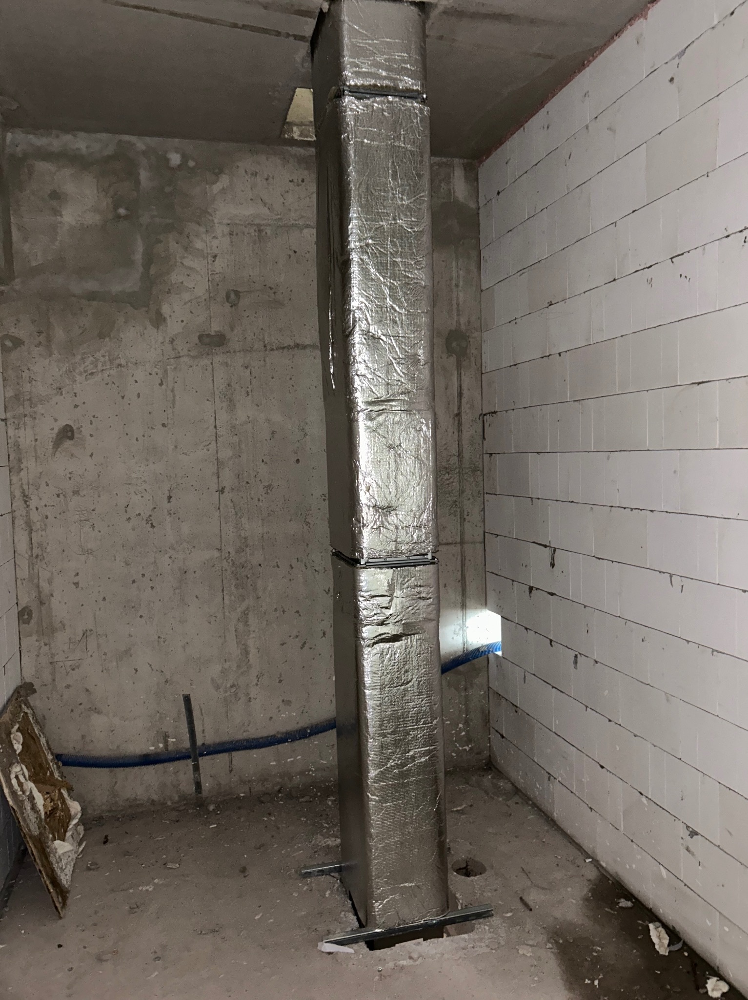 Pionowy kanał wentylacyjny owinięty folią izolacyjną, zamontowany w surowym pomieszczeniu z betonowymi i bloczkowymi ścianami, widoczne elementy instalacji i resztki materiałów budowlanych.