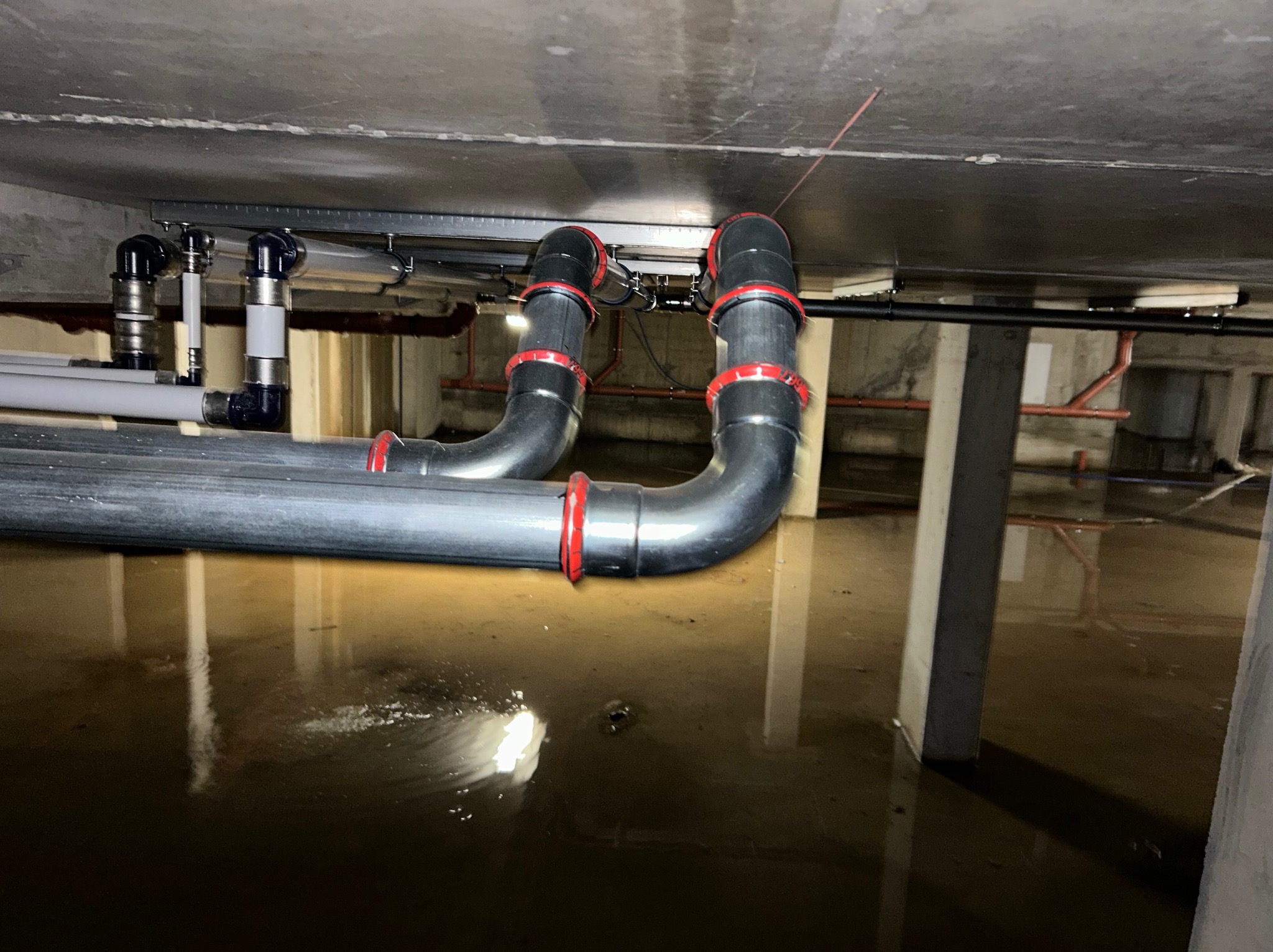 Instalacja rur kanalizacyjnych pod sufitem piwnicy, z widoczną wodą na podłodze.