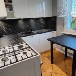 Nowoczesna kuchnia z białymi szafkami, czarnym blatem imitującym marmur i płytą gazową, oświetlenie punktowe pod szafkami górnymi, widok na stół z blatem w tym samym stylu.