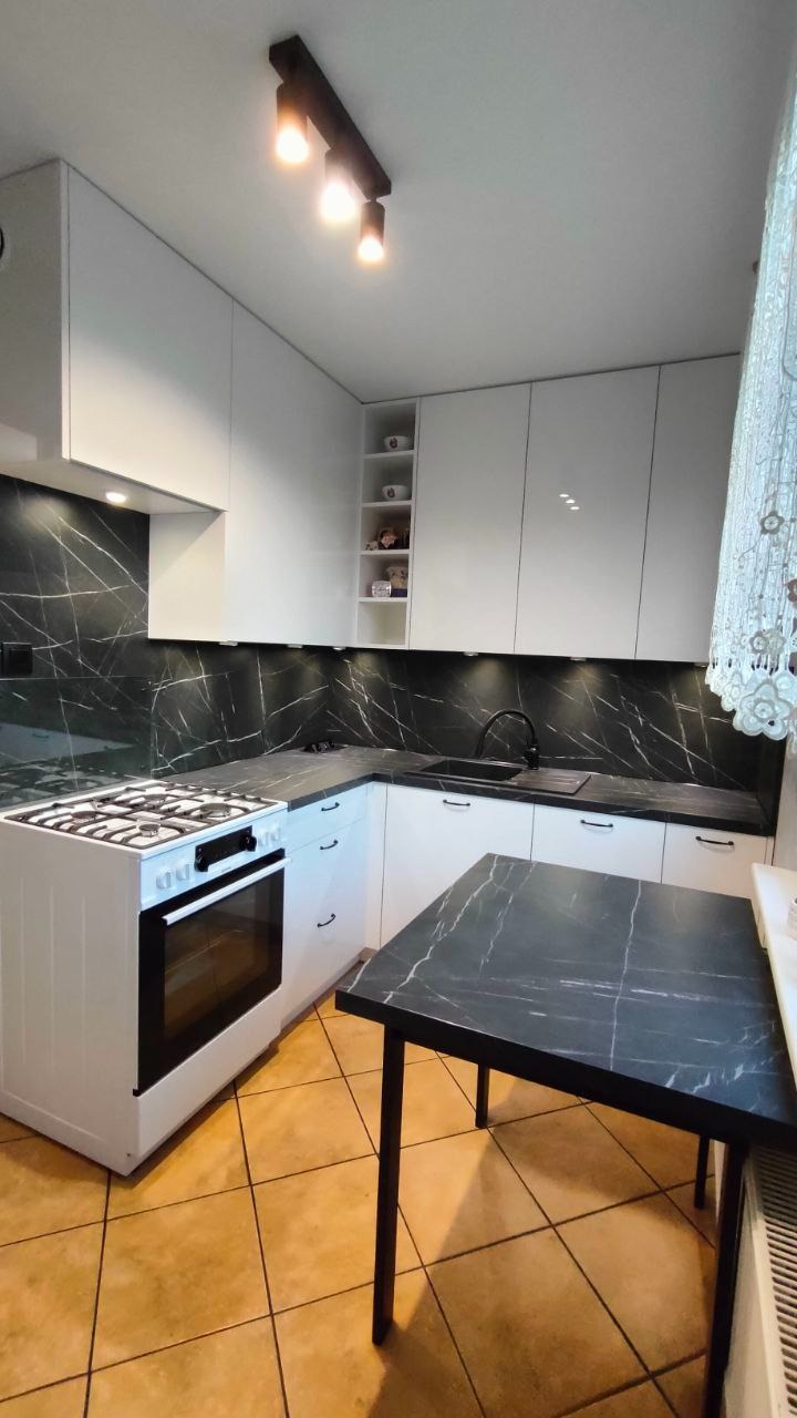 Nowoczesna biała kuchnia z czarnym blatem imitującym marmur i białym piekarnikiem, oświetlona punktowymi lampami sufitowymi i podszafkowymi, z widocznym fragmentem stołu z blatem w tym samym stylu.