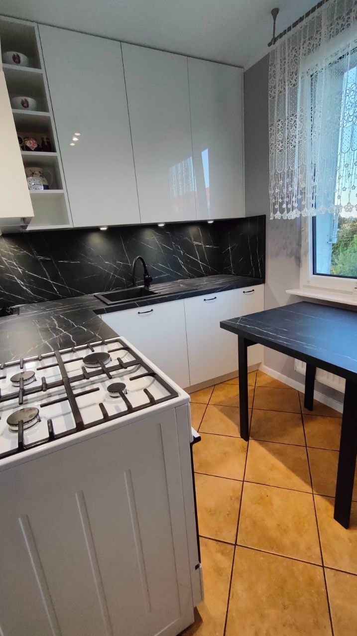 Nowoczesna kuchnia z białymi szafkami, czarnym blatem imitującym marmur i płytą gazową, oświetlenie punktowe pod szafkami górnymi, widok na stół z blatem w tym samym stylu.