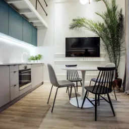 Nowoczesna kuchnia z szarymi szafkami, marmurowym backsplashiem, okrągłym białym stołem i krzesłami w stylu skandynawskim, z dużym telewizorem na ścianie i rośliną doniczkową.