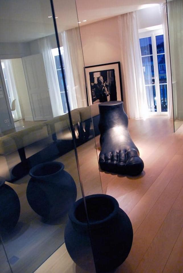 Nowoczesny salon z drewnianą podłogą, szklaną ścianą, czarnymi wazonami i rzeźbą stopy, oświetlony naturalnym światłem wpadającym przez okno z białymi zasłonami.