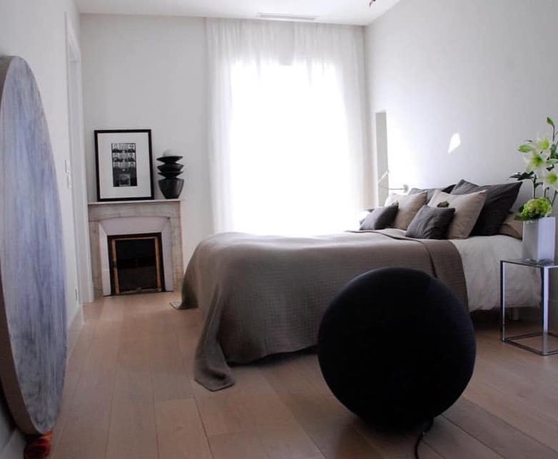 Jasna sypialnia z drewnianą podłogą, minimalistycznym kominkiem z czarnym wkładem i dekoracyjną czarną kulą na podłodze, doświetlona przez duże okno z białymi zasłonami.