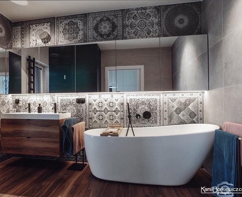 Nowoczesna łazienka z drewnianą podłogą, wolnostojącą białą wanną, umywalką nablatową z drewnianą szafką i lustrami z dekoracyjnymi płytkami w stylu orientalnym.