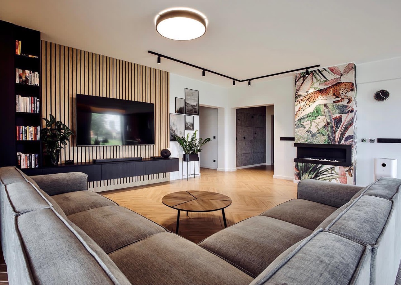Nowoczesny salon z drewnianą ścianą, telewizorem, szarą sofą, okrągłym stolikiem kawowym i kominkiem z motywem dżungli.
