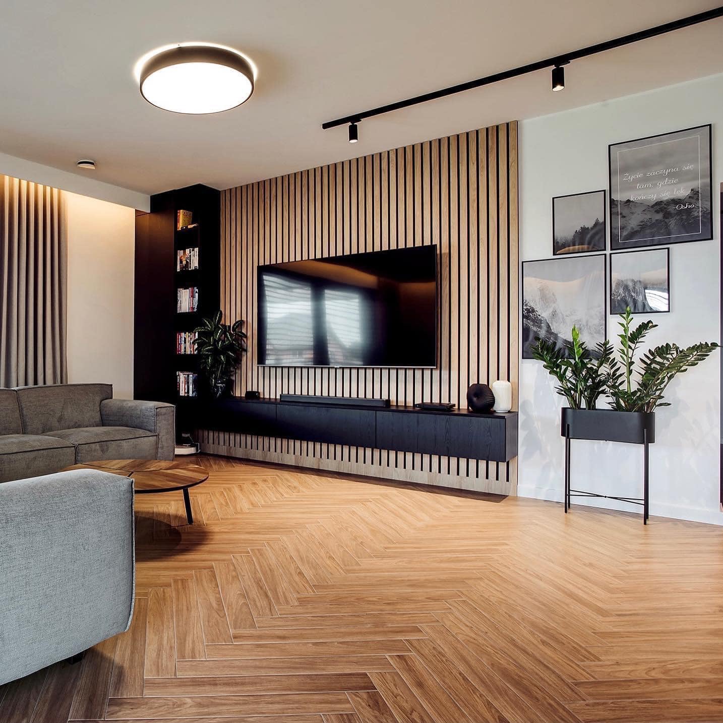 Nowoczesny salon z drewnianą ścianą z lameli, telewizorem na czarnej szafce RTV, parkietem w jodełkę, szarą sofą i roślinami doniczkowymi.