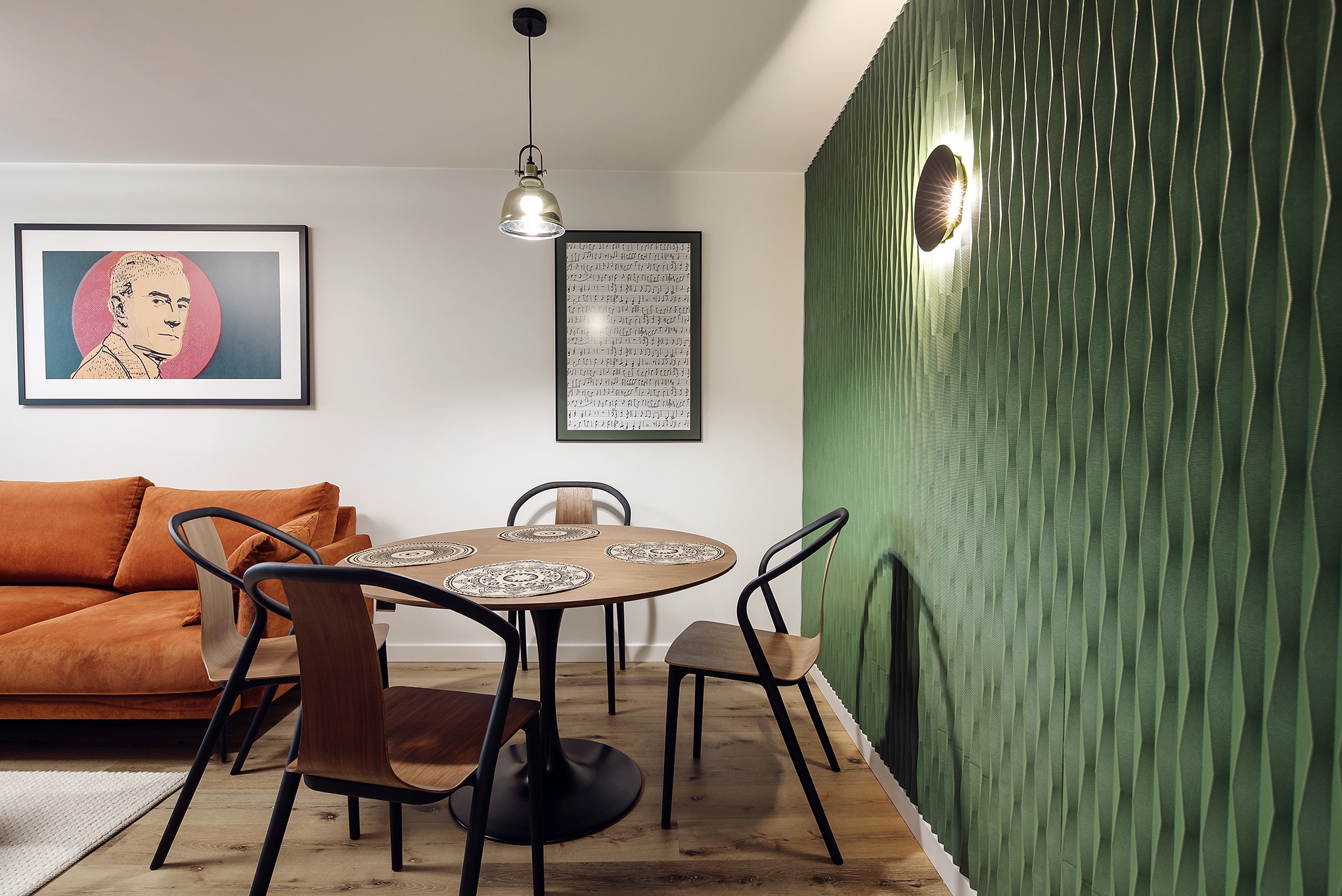 Nowoczesna jadalnia z okrągłym stołem na czarnej nodze, drewnianymi krzesłami z czarnymi nogami, pomarańczową sofą w tle i zieloną ścianą z oświetleniem punktowym.
