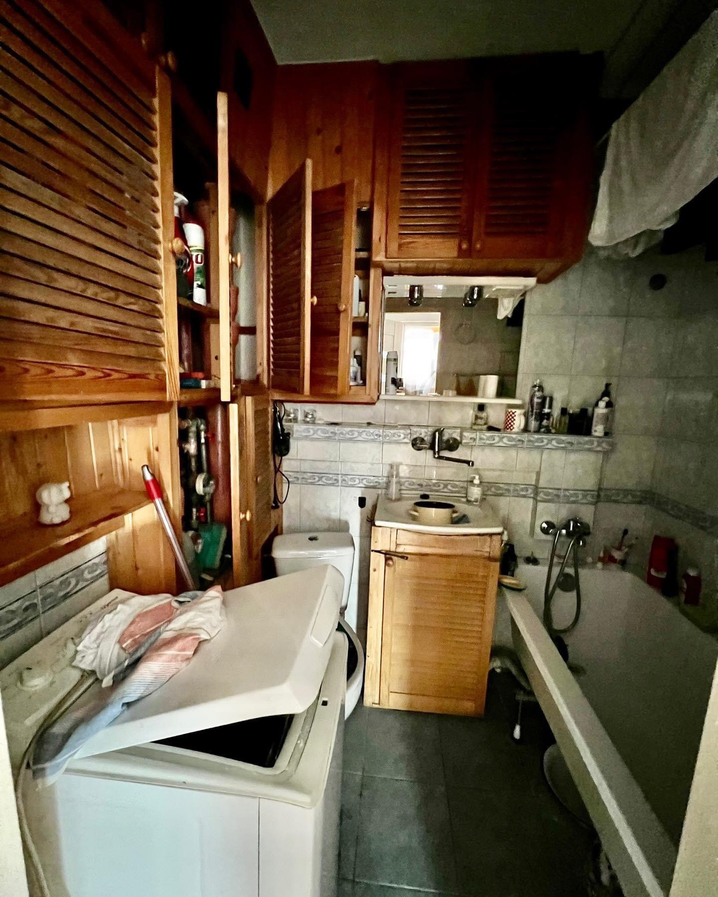 Wnętrze łazienki z drewnianymi szafkami, pralką z otwartą klapą i wanną, widać elementy instalacji hydraulicznej i różnorodne przedmioty na półkach.