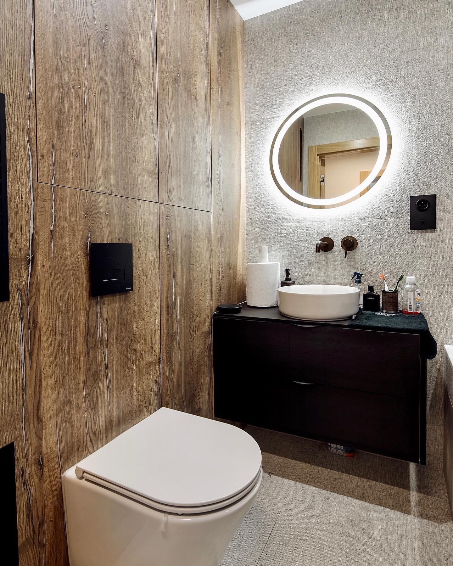 Nowoczesna łazienka z drewnianą zabudową, okrągłym lustrem z podświetleniem LED, białą umywalką nablatową i wiszącą toaletą.