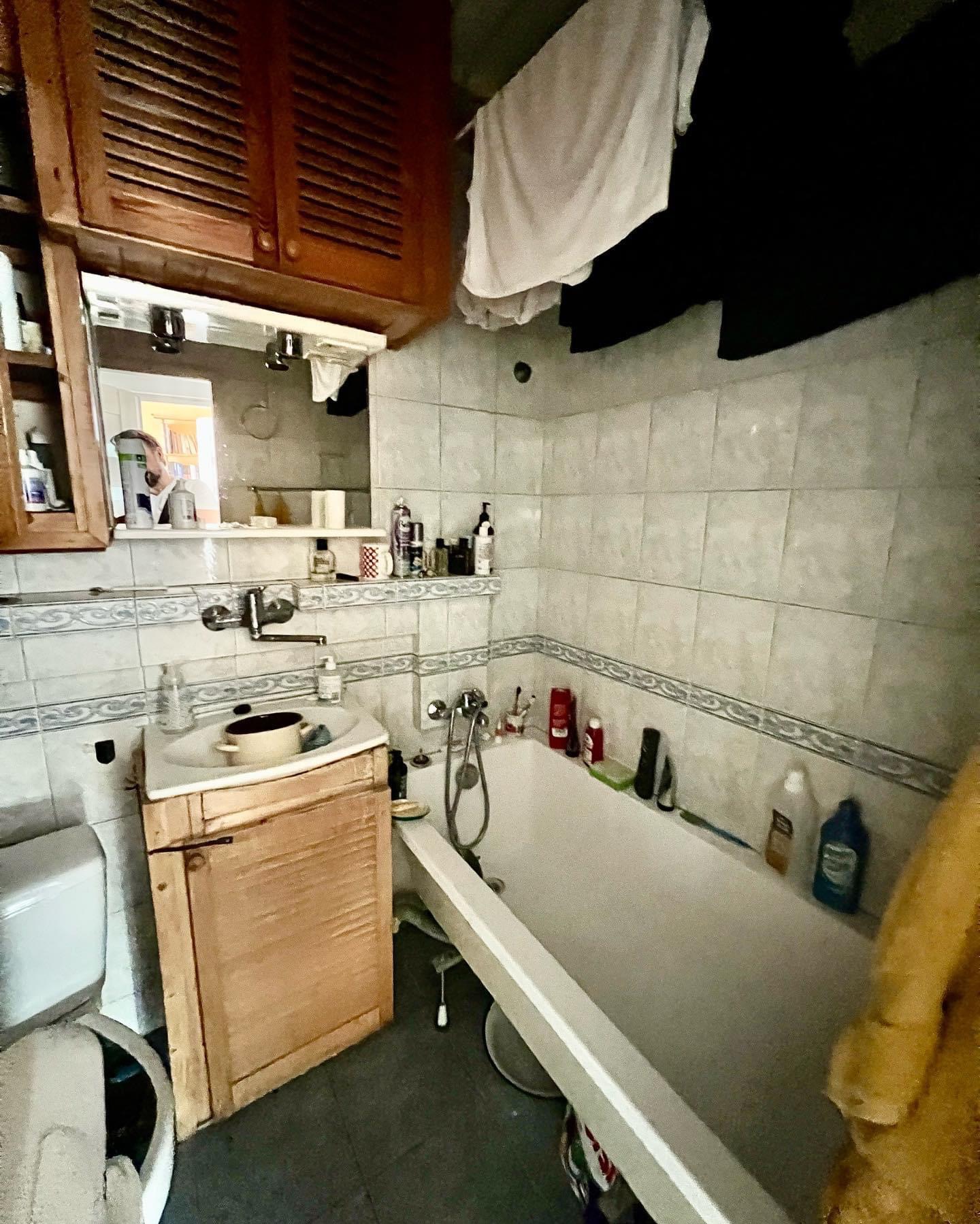 Wnętrze łazienki z wanną, umywalką z szafką z drewnianymi lamelami, lustrem i kafelkami w odcieniach szarości, na których wiszą ubrania.