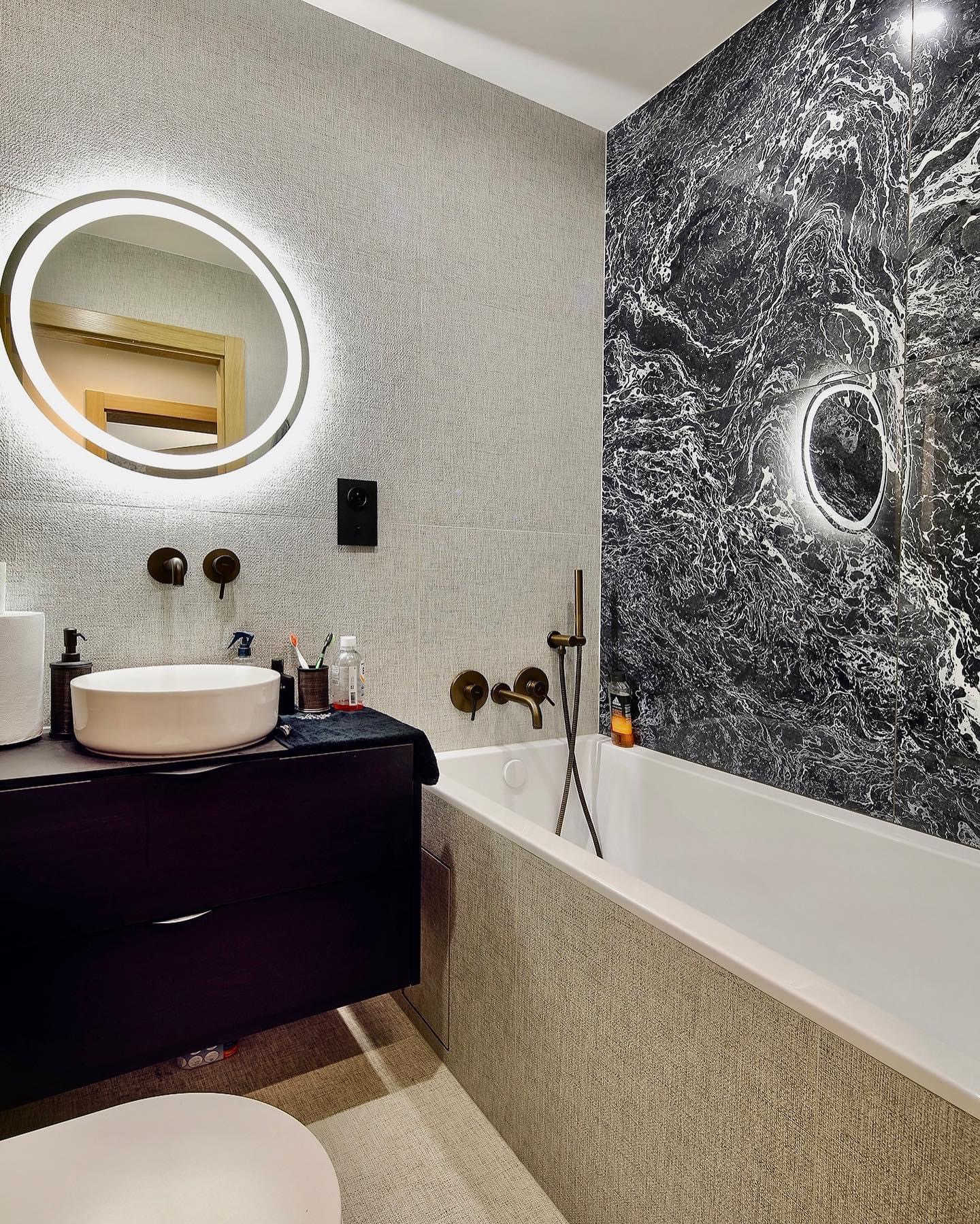 Nowoczesna łazienka z okrągłym lustrem podświetlanym LED, białą umywalką nablatową, ciemną szafką pod umywalką, wanną obudowaną płytkami imitującymi tkaninę i ścianą wyłożoną czarnym marmurem...