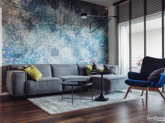 Szary narożnik z żółtą poduszką na tle ściany pokrytej abstrakcyjnym wzorem w odcieniach błękitu, granatu i szarości; obok okno z żaluzjami, czarny stolik kawowy i fotel w kolorze granatowym.