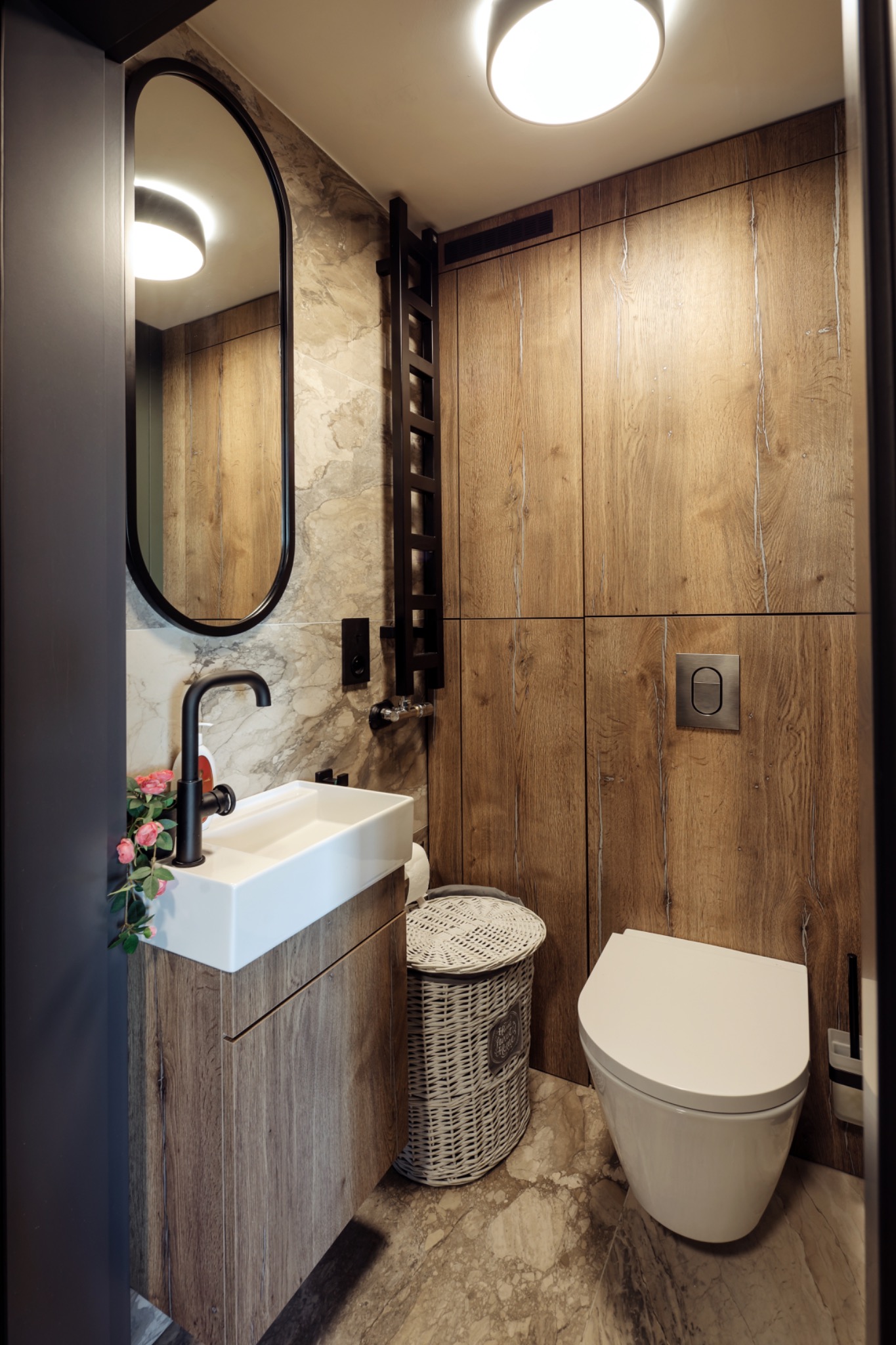 Nowoczesna łazienka z drewnianymi akcentami: umywalka nablatowa, czarna bateria, owalne lustro w czarnej ramie, grzejnik drabinkowy i toaleta podwieszana na tle drewnianej zabudowy.