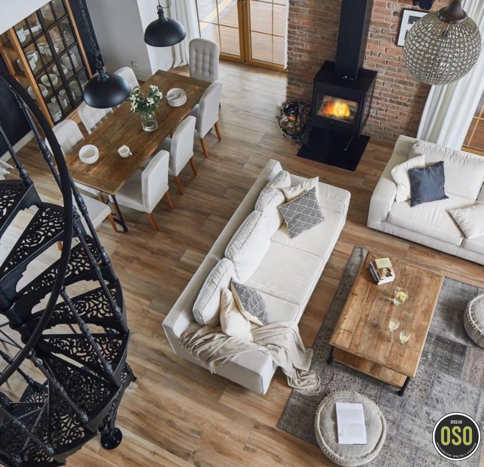 Widok z góry na salon z drewnianym stołem jadalnym, białymi krzesłami, kominkiem, białymi sofami, drewnianym stolikiem kawowym i czarnymi kręconymi schodami.