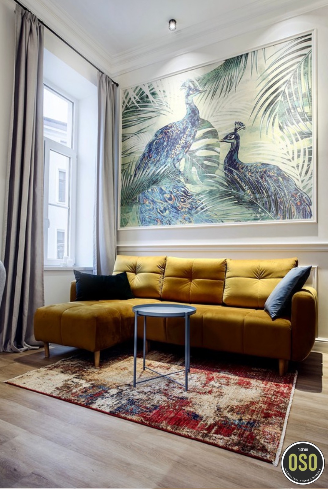 Żółta, welurowa sofa narożna z czarną i niebieską poduszką na tle ściany z obrazem przedstawiającym pawie i tropikalne liście; mały, okrągły, metalowy stolik kawowy oraz wzorzysty dywan...