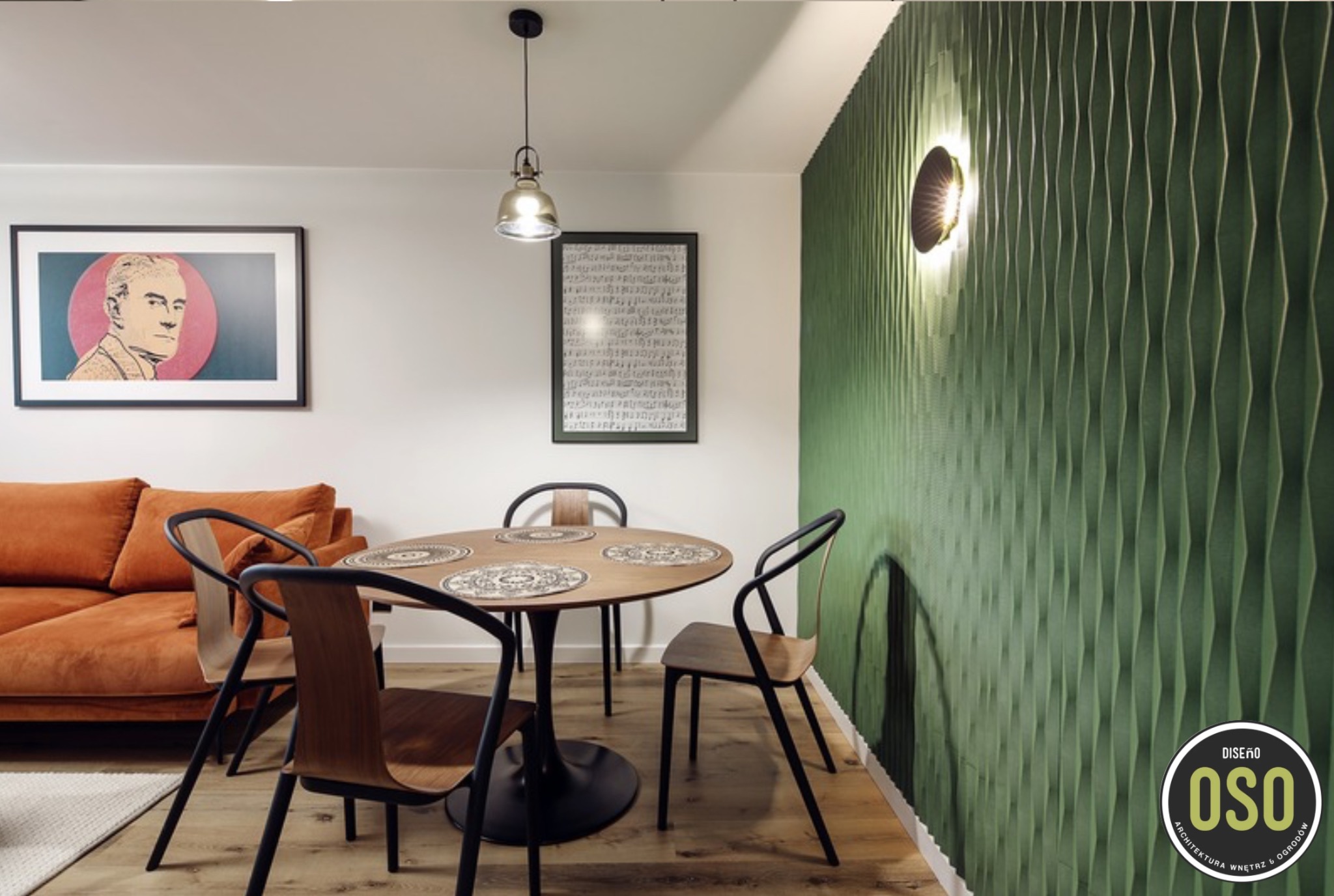 Nowoczesna jadalnia z okrągłym stołem na tulipanowej nodze, drewnianymi krzesłami o giętych oparciach, pomarańczową sofą w tle i zieloną ścianą z trójwymiarową fakturą oświetloną okrągłym kinkietem.