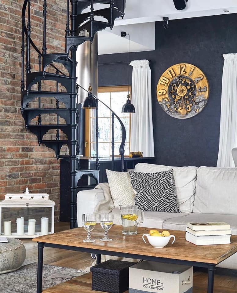 Salon w stylu industrialnym z ceglaną ścianą, spiralnymi schodami, dekoracyjnym zegarem na ciemnej ścianie i drewnianym stolikiem kawowym z cytrynową wodą.