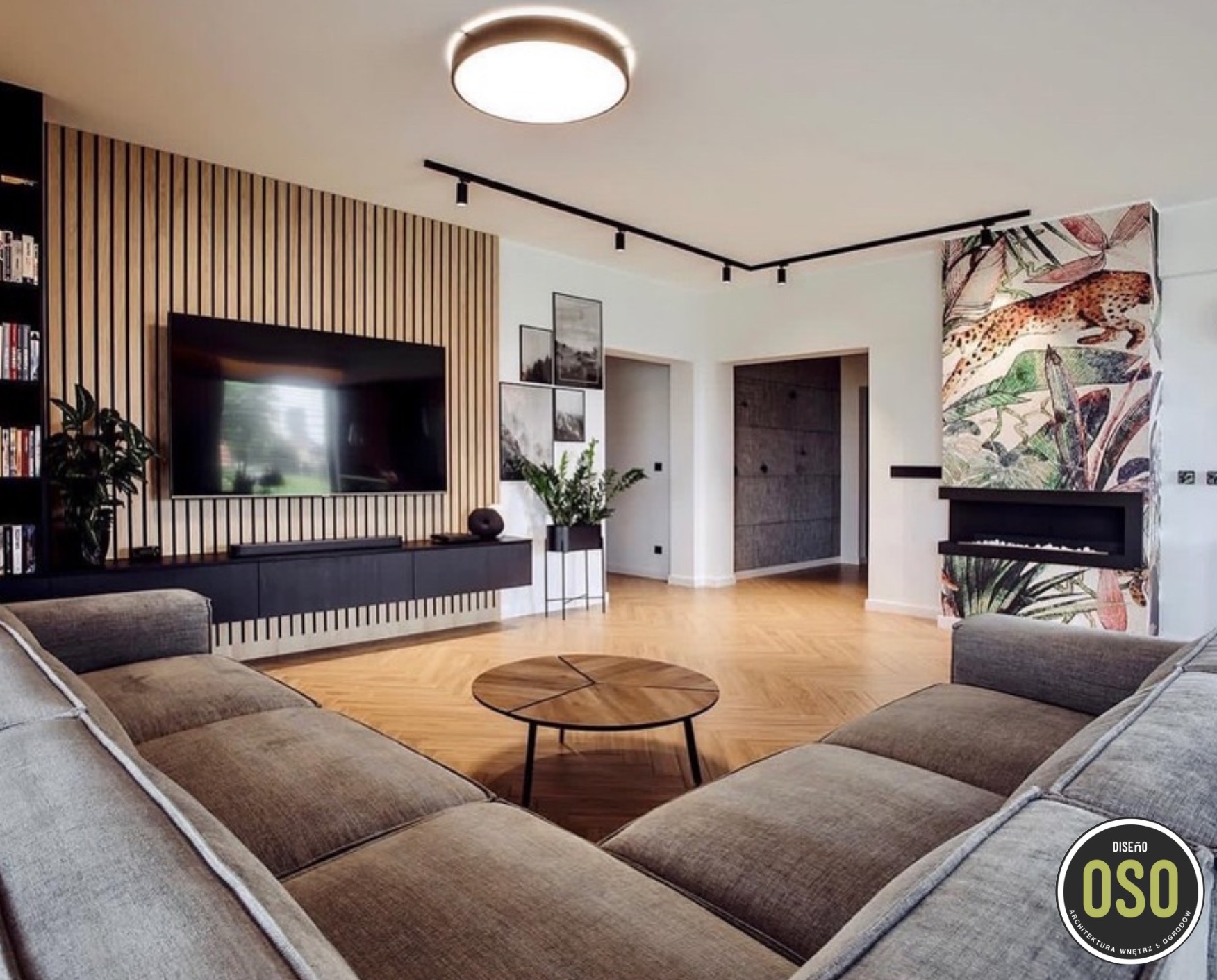 Nowoczesny salon z drewnianą ścianą z lamelami, telewizorem, niską szafką RTV, okrągłym stolikiem kawowym i narożną sofą w odcieniach szarości, parkiet w jodełkę, oświetlenie szynowe i dekoracyjny...