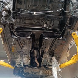 BREMS - Podwozie samochodu zabezpieczone folią i podniesione na żółtym podnośniku, po nałożeniu czarnej powłoki ochronnej.