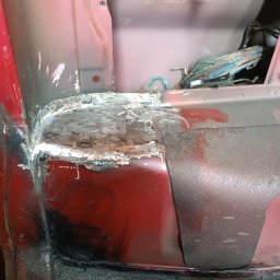 BREMS - Fragment karoserii samochodu w kolorze czerwonym, z widocznymi śladami napraw blacharskich i spawania, odkryta wiązka przewodów w tle.