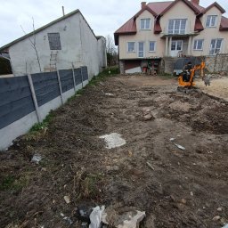 BREMS - Pomarańczowa mini-koparka na nierównym, przygotowanym terenie budowy z dwoma budynkami w tle, widoczne ślady opon i resztki materiałów budowlanych.