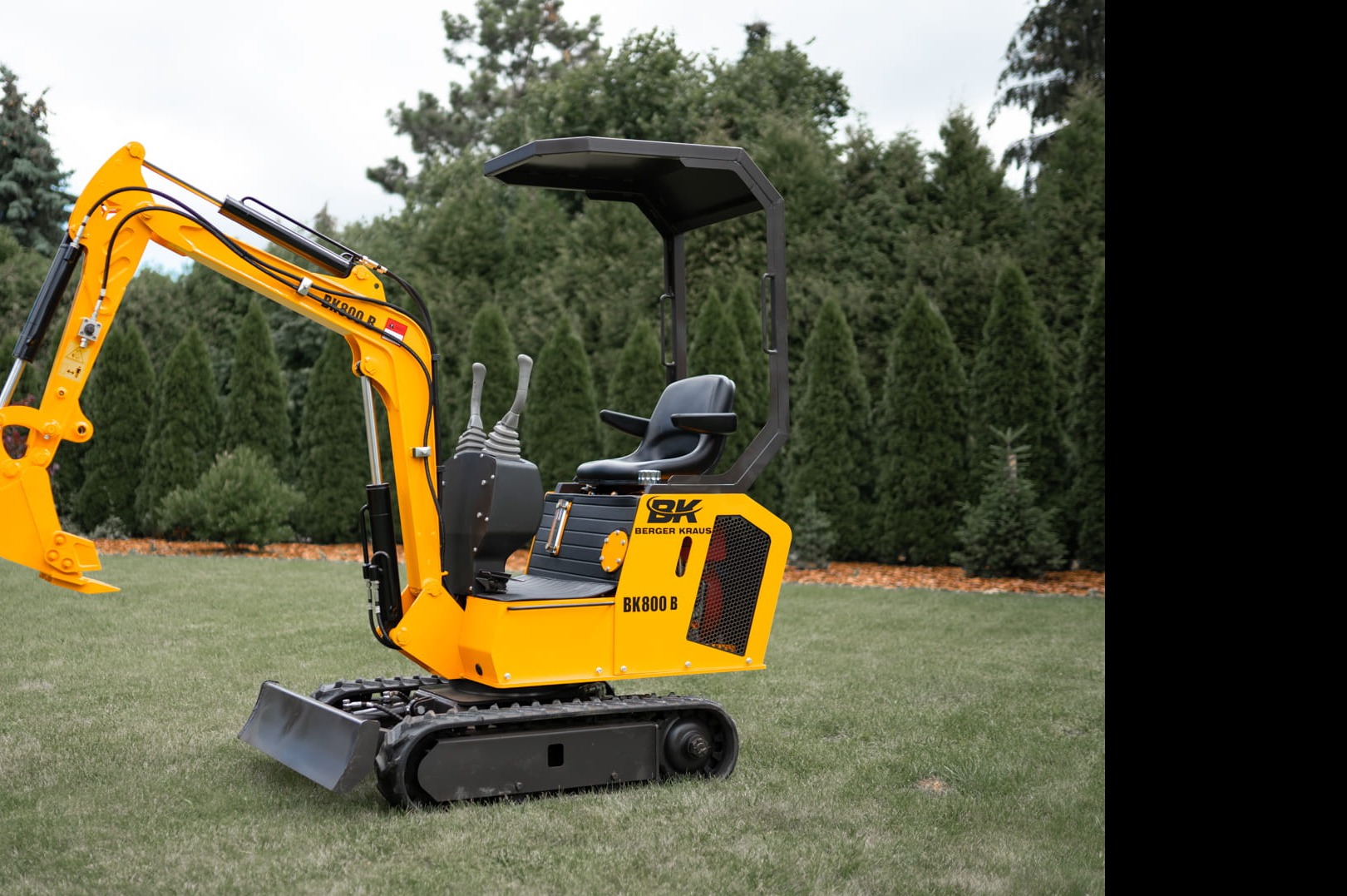 Żółto-czarna mini koparka Berger Kraus BK800 B na trawniku przed rzędem wysokich, zielonych iglaków.