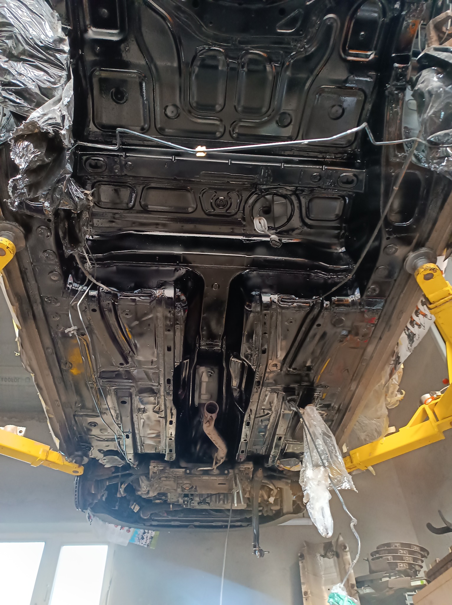 Podwozie samochodu zabezpieczone folią i podniesione na żółtym podnośniku, po nałożeniu czarnej powłoki ochronnej.